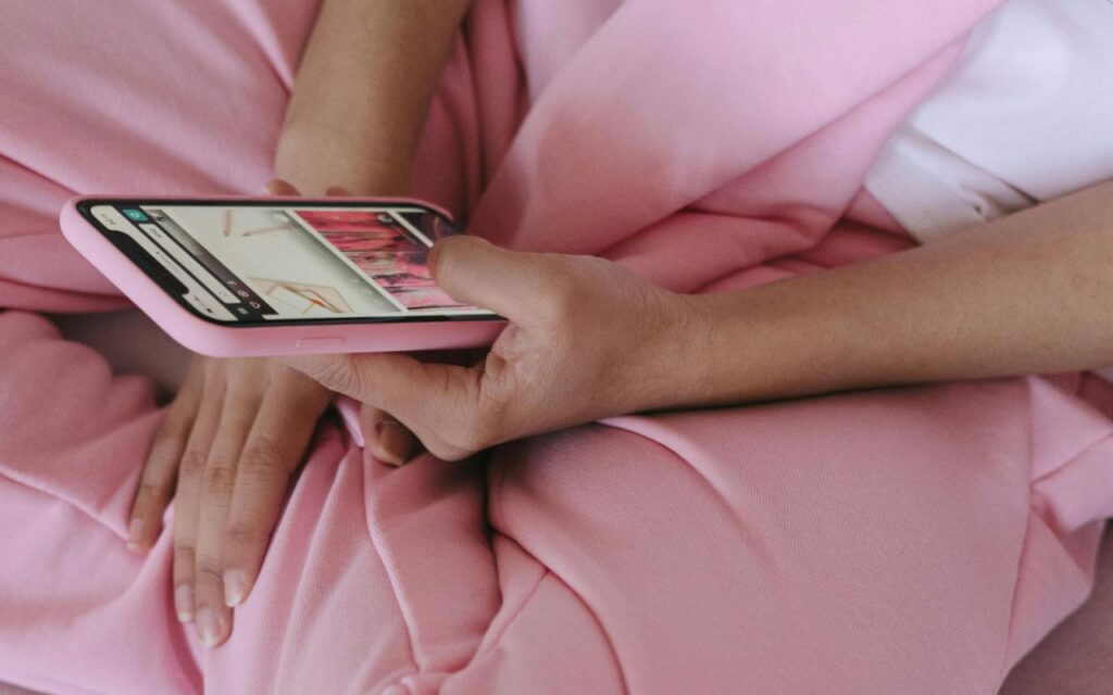 Žena u roze trenerci i beloj majici sedi na kauči i gleda u telefon